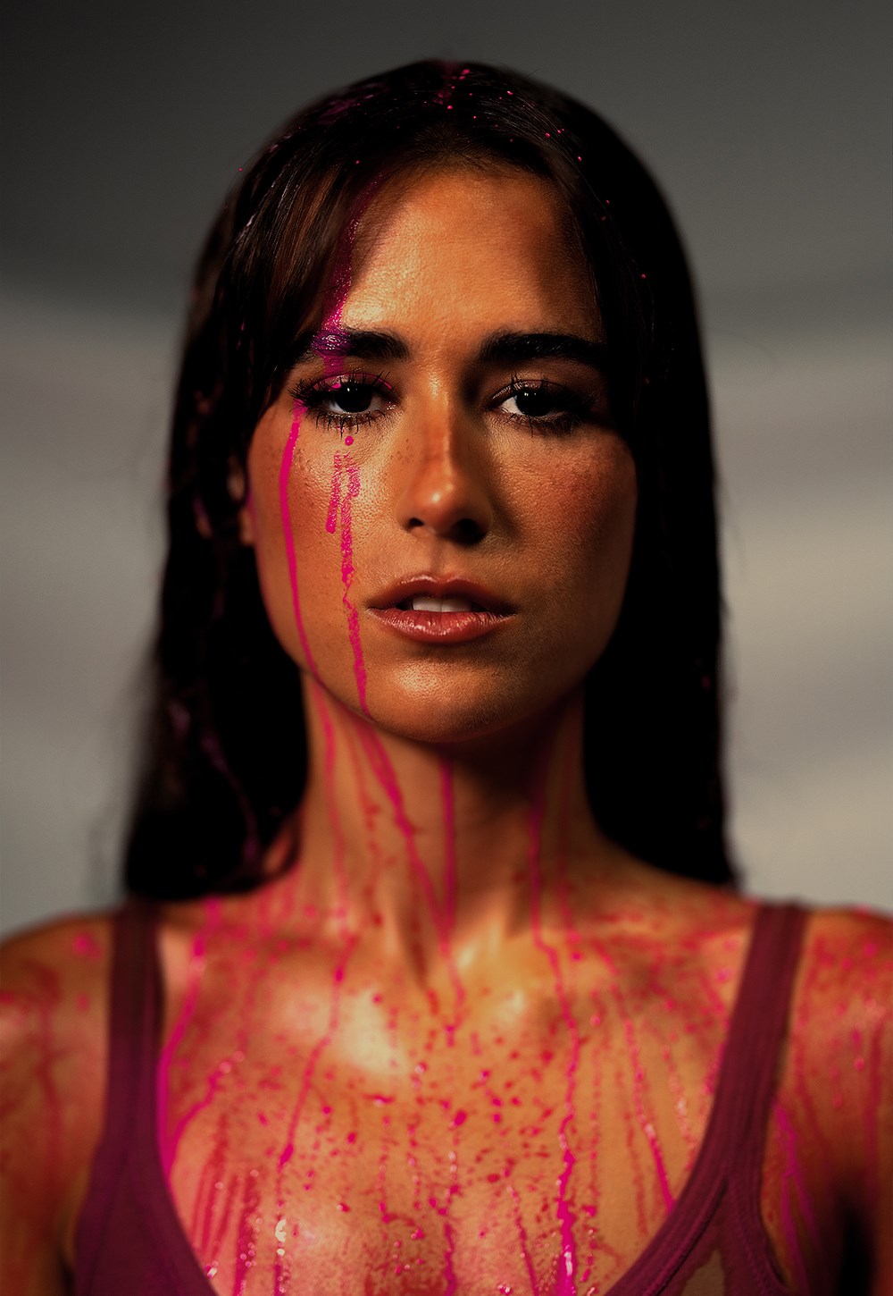 Portrait of a woman with hot-pink paint dripping down her face and chest, wearing a burgundy top, moody lighting.