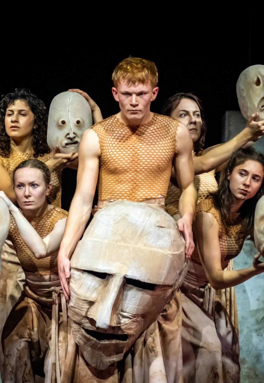 A male performer wearing a tan mesh top stands at center, surrounded by others in beige outfits, holding a large paper-mache head prop.